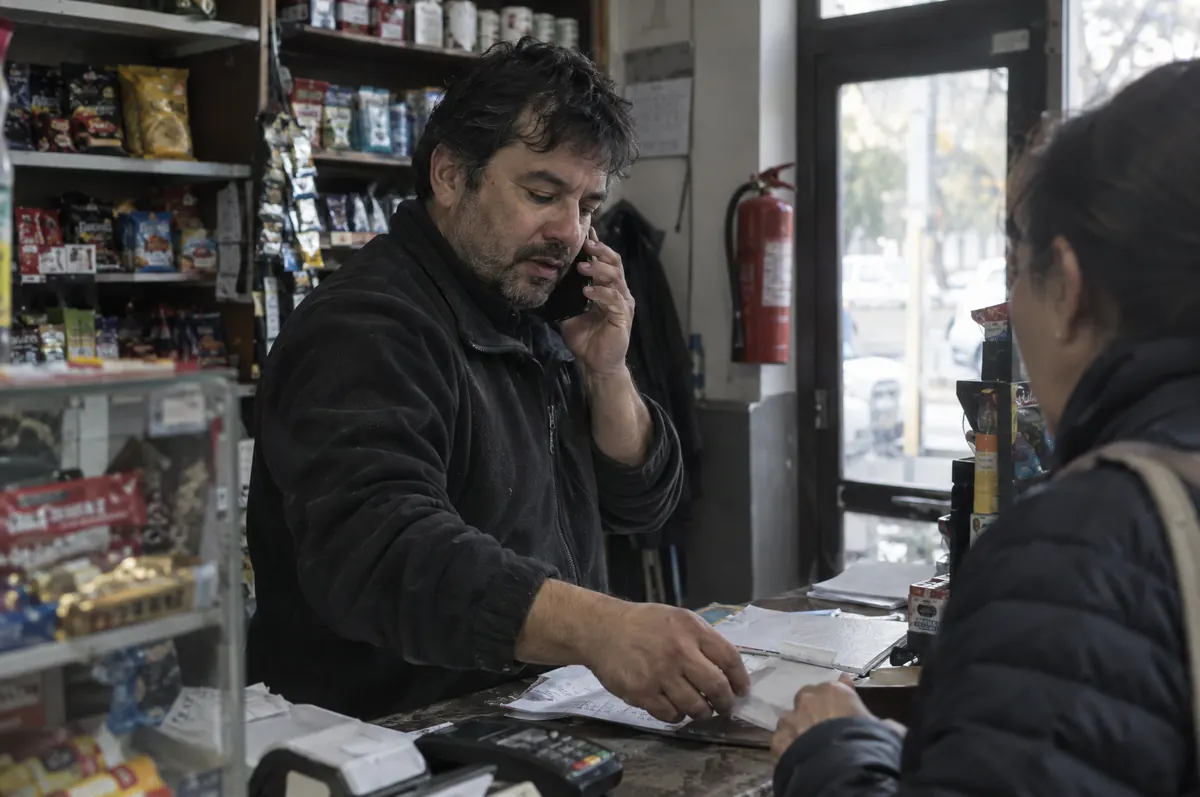 Dueño de un minimarket atendiendo a una clienta mientras habla por teléfono, con un extintor en la pared